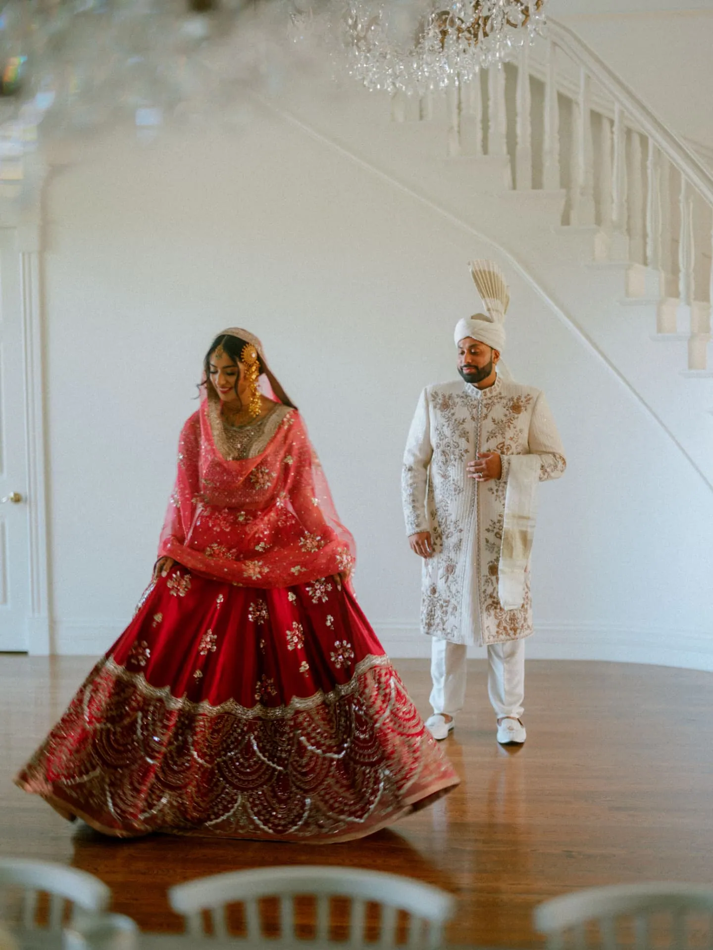 Bride in a red lehenga turning as the groom watches in a white hall
