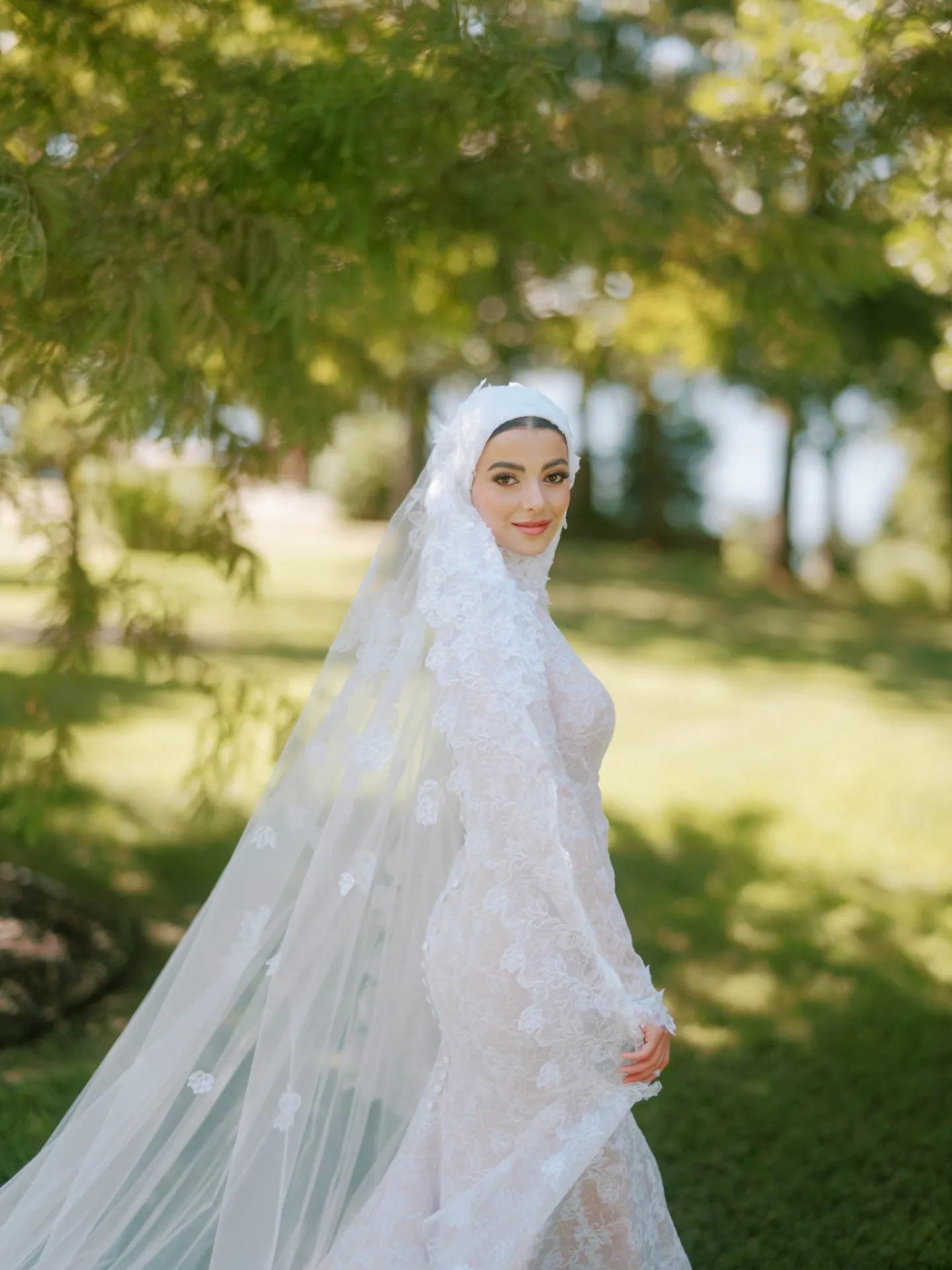 Bride in a lace gown and veil beneath green trees