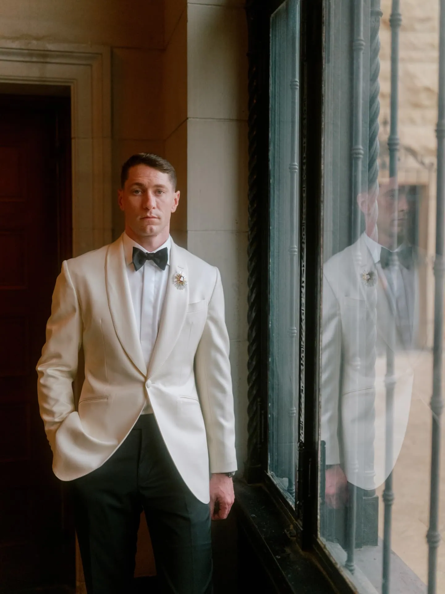 Groom in a white dinner jacket reflected in a tall window