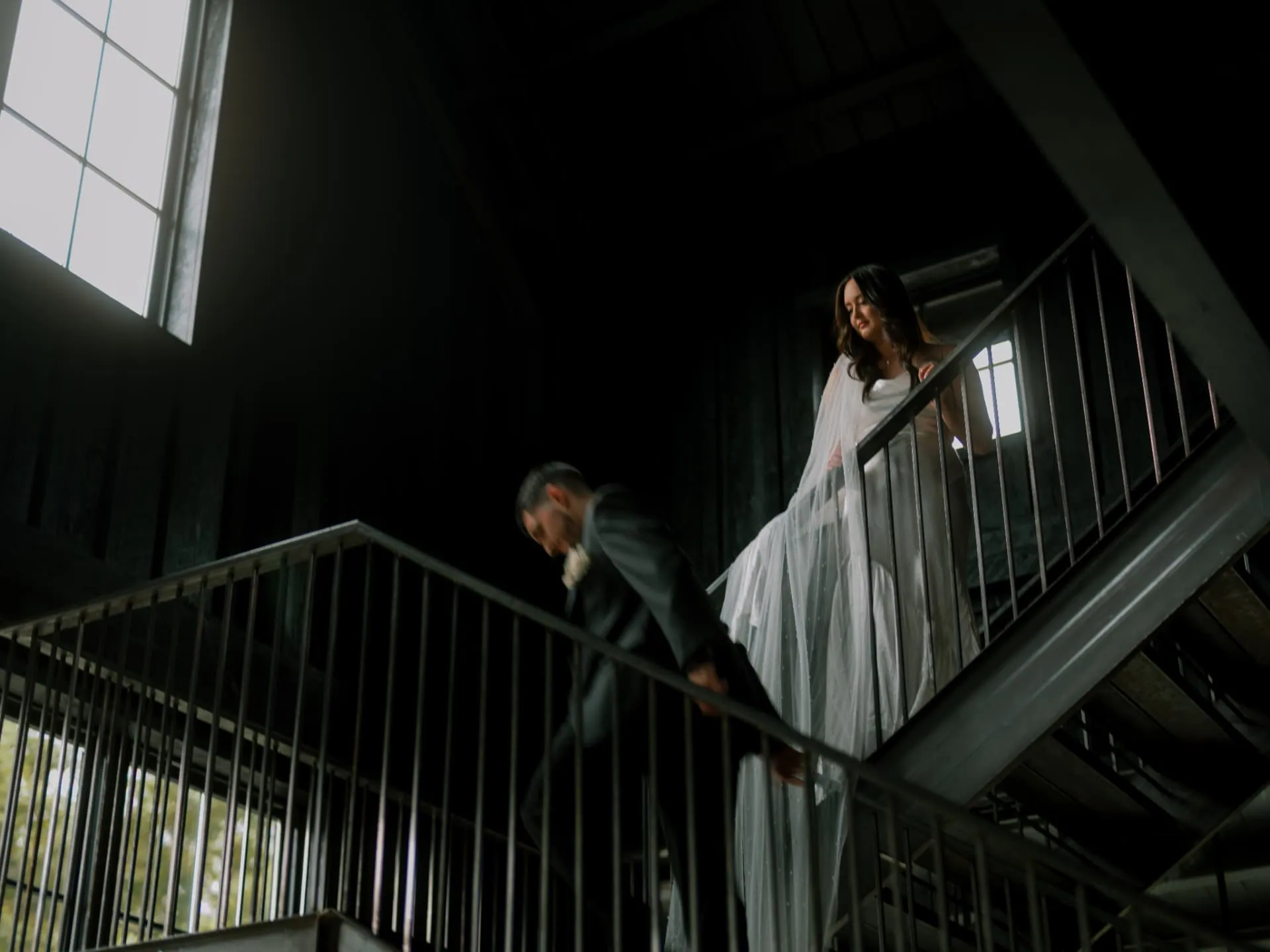 Bride and groom on a dark stairwell lit by a single window