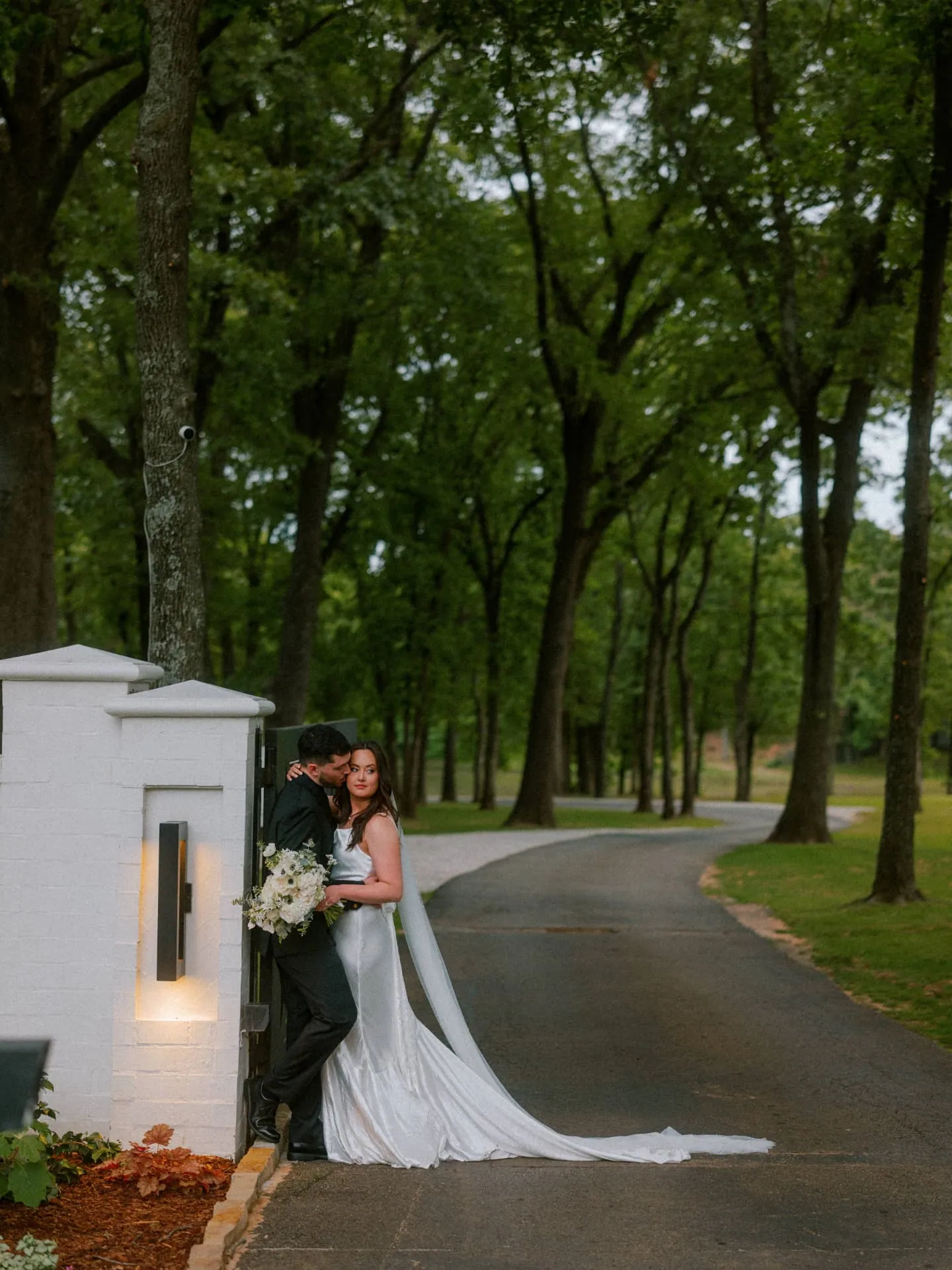 Couple leaning against a white gate on a tree-lined drive